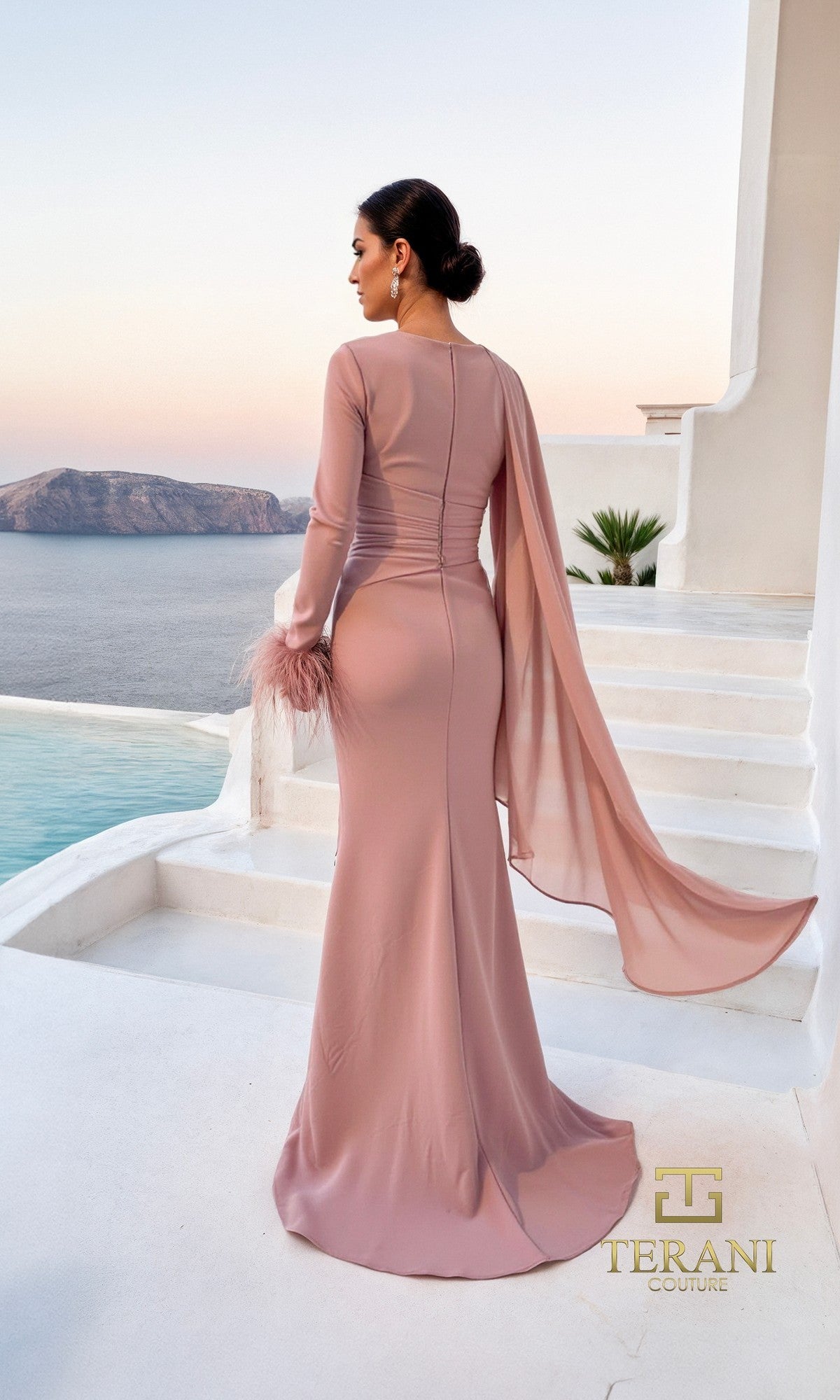 A woman in the Terani 261M6403 long formal dress, featuring long sleeves and a sheer cape, stands on white steps overlooking coastal cliffs and water.